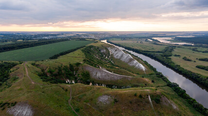 Panoramic aerial view over the top of a summer landscape of a green agricultural field, hills or mountains, a large river, and a forest belt at sunset, captured from a helicopter as a bird's eye view