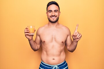 Fototapeta premium Young handsome man with beard on vacation wearing swimwear drinking glass of orange juice smiling with an idea or question pointing finger with happy face, number one