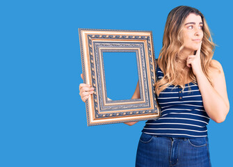 Young caucasian woman holding empty frame serious face thinking about question with hand on chin,...