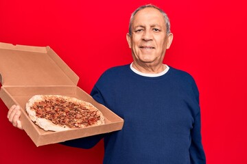 Senior man with grey hair holding delivery cardboard of italian pizza looking positive and happy standing and smiling with a confident smile showing teeth