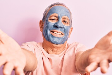 Senior man with grey hair wearing mud mask looking at the camera smiling with open arms for hug. cheerful expression embracing happiness.