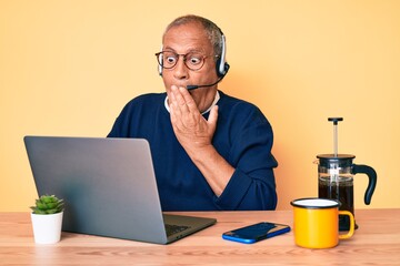 Senior handsome man with gray hair working at the office wearing operator headset covering mouth with hand, shocked and afraid for mistake. surprised expression