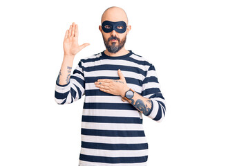 Naklejka premium Young handsome man wearing burglar mask swearing with hand on chest and open palm, making a loyalty promise oath