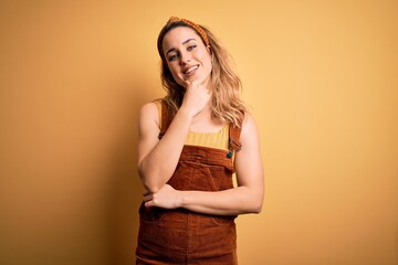 Obraz premium Young beautiful blonde woman wearing overalls and diadem standing over yellow background looking confident at the camera smiling with crossed arms and hand raised on chin. Thinking positive.