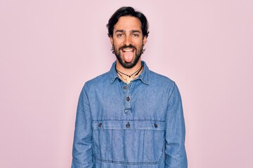Young handsome hispanic bohemian man wearing hippie style over pink background sticking tongue out happy with funny expression. Emotion concept.