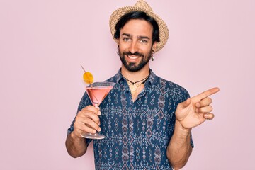Young handsome hispanic bohemian hipster man with bear drinking a summer cocktail very happy pointing with hand and finger to the side