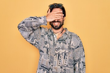 Young handsome hispanic bohemian man wearing hippie style and sunglasses smiling and laughing with hand on face covering eyes for surprise. Blind concept.