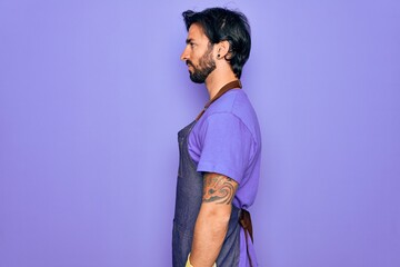 Young handsome hispanic clenaer man wearing housework apron and washing gloves looking to side, relax profile pose with natural face with confident smile.