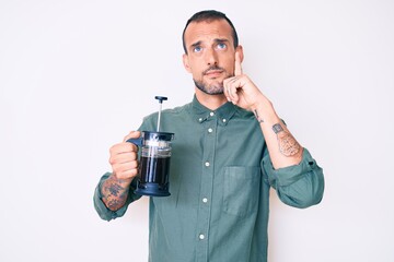 Young handsome man with tattoo holding french coffee maker serious face thinking about question...