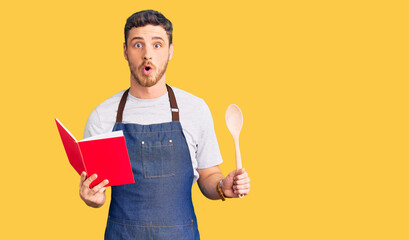 Handsome young man with bear wearing professional baker apron reading cooking recipe book scared...