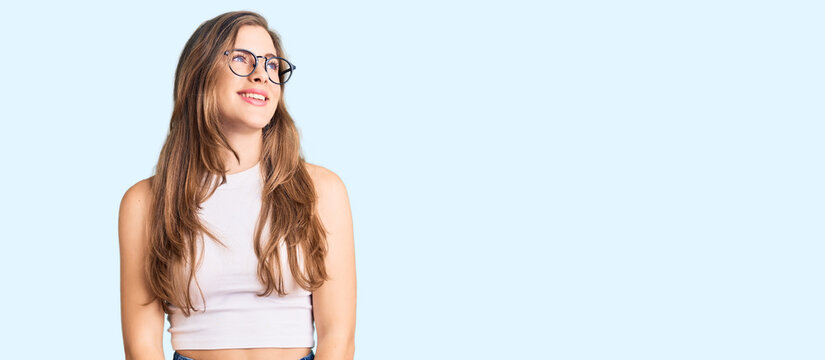 Beautiful caucasian young woman wearing casual clothes and glasses looking away to side with smile on face, natural expression. laughing confident.