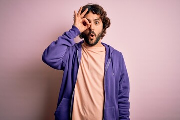 Young handsome sporty man with beard wearing casual sweatshirt over pink background doing ok gesture shocked with surprised face, eye looking through fingers. Unbelieving expression.