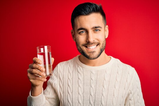 Young handsome man smiling happy and confident. Standing with smile on face holding glass of healthy water to refreshment over isolated red background