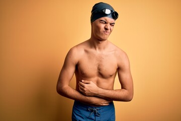 Young handsome man shirtless wearing swimsuit and swim cap over isolated yellow background with hand on stomach because indigestion, painful illness feeling unwell. Ache concept.