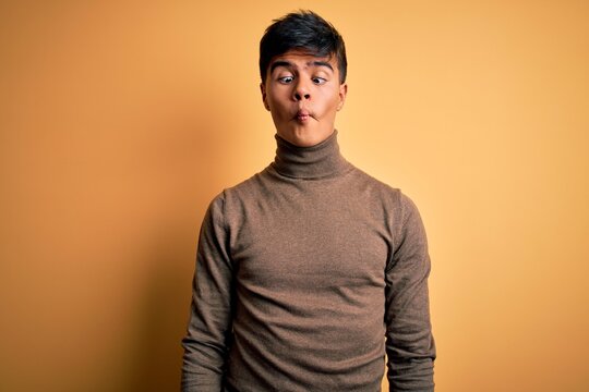 Young handsome man wearing casual turtleneck sweater over isolated yellow background making fish face with lips, crazy and comical gesture. Funny expression.
