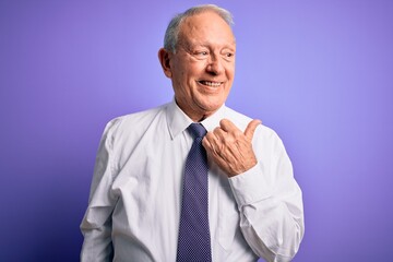 Grey haired senior business elegant man standing over purple isolated background smiling with happy face looking and pointing to the side with thumb up.
