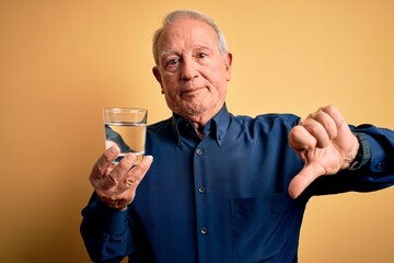 Grey haired senior man drinking a fresh glass of water over yellow isolated background with angry face, negative sign showing dislike with thumbs down, rejection concept