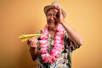 Grey haired senior man wearing summer hat and hawaiian lei holding paper plane on vacation with happy face smiling doing ok sign with hand on eye looking through fingers