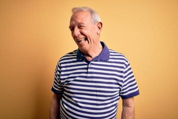 Grey haired senior man wearing casual navy striped t-shirt standing over yellow background winking...
