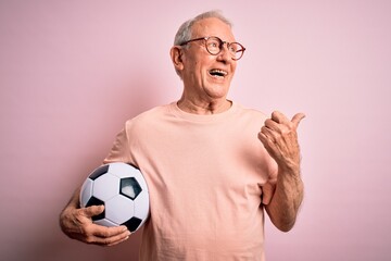 Grey haired senior football player man holding soccer ball over pink isolated background pointing and showing with thumb up to the side with happy face smiling