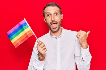 Young handsome man holding rainbow lgbtq flag pointing thumb up to the side smiling happy with open mouth