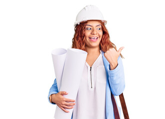 Young latin woman wearing architect hardhat and leather bag holding blueprints pointing thumb up to the side smiling happy with open mouth