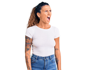 Fototapeta premium Young hispanic woman with tattoo wearing casual white tshirt angry and mad screaming frustrated and furious, shouting with anger. rage and aggressive concept.