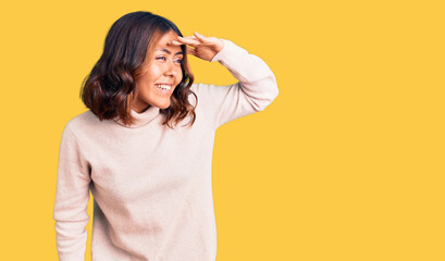 Young beautiful mixed race woman wearing winter turtleneck sweater very happy and smiling looking far away with hand over head. searching concept.