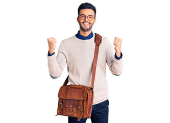 Young handsome hispanic man wearing leather bag screaming proud, celebrating victory and success very excited with raised arms