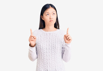Young beautiful chinese woman wearing casual sweater pointing up looking sad and upset, indicating...