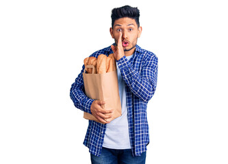 Handsome latin american young man holding paper bag with bread hand on mouth telling secret rumor, whispering malicious talk conversation