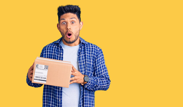 Handsome latin american young man holding delivery package scared and amazed with open mouth for surprise, disbelief face