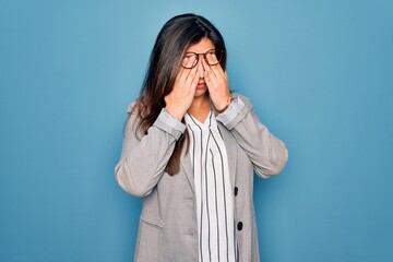Young hispanic business woman wearing glasses standing over blue isolated background rubbing eyes...