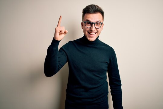 Young handsome caucasian man wearing glasses and casual sweater over isolated background pointing finger up with successful idea. Exited and happy. Number one.