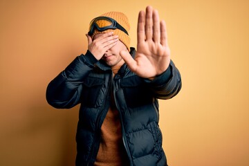 Young handsome caucasian man wearing hat, coat and ski glasses for winter and snow weather covering eyes with hands and doing stop gesture with sad and fear expression. Embarrassed and negative