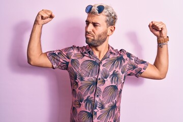 Handsome blond man on vacation wearing casual shirt and sunglasses over pink background showing arms muscles smiling proud. Fitness concept.