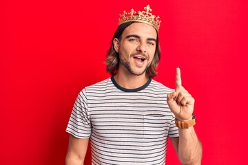 Young handsome man wearing prince crown smiling with an idea or question pointing finger up with...