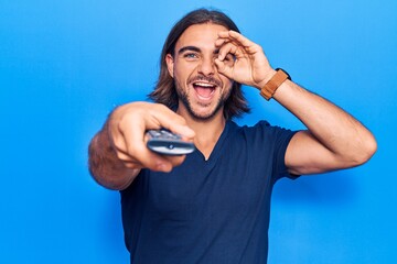 Naklejka premium Young handsome man holding television remote control smiling happy doing ok sign with hand on eye looking through fingers