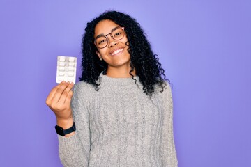 Young african american curly woman holding pills standing over isolated purple background with a happy face standing and smiling with a confident smile showing teeth