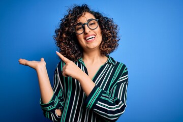 Young beautiful curly arab woman wearing striped shirt and glasses over blue background amazed and smiling to the camera while presenting with hand and pointing with finger.