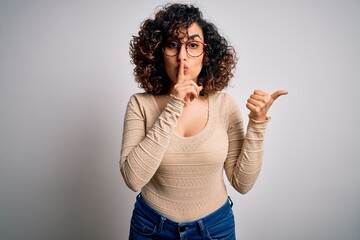 Young beautiful curly arab woman wearing casual t-shirt and glasses over white background asking to be quiet with finger on lips pointing with hand to the side. Silence and secret concept.