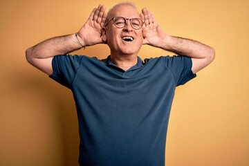 Fototapeta na wymiar Middle age handsome hoary man wearing casual polo and glasses over yellow background Smiling cheerful playing peek a boo with hands showing face. Surprised and exited
