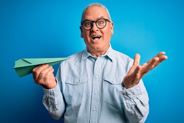 Middle age handsome hoary man holding paper airplane over isolated blue background very happy and excited, winner expression celebrating victory screaming with big smile and raised hands