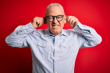 Middle age handsome hoary man wearing casual striped shirt and glasses over red background Smiling pulling ears with fingers, funny gesture. Audition problem