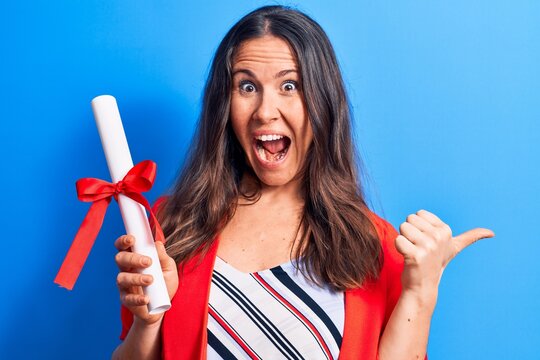 Young beautiful brunette smart woman holding graduated degree diploma over blue background pointing thumb up to the side smiling happy with open mouth