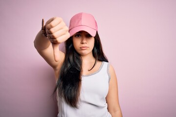 Obraz na płótnie Canvas Young brunette woman wearing casual sport cap over pink background looking unhappy and angry showing rejection and negative with thumbs down gesture. Bad expression.