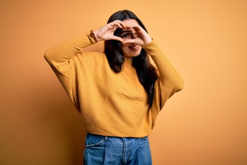 Young brunette woman wearing glasses and casual sweater over yellow isolated background Doing heart shape with hand and fingers smiling looking through sign