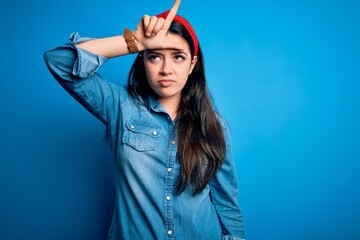 Young brunette woman wearing casual denim shirt over blue isolated background making fun of people with fingers on forehead doing loser gesture mocking and insulting.