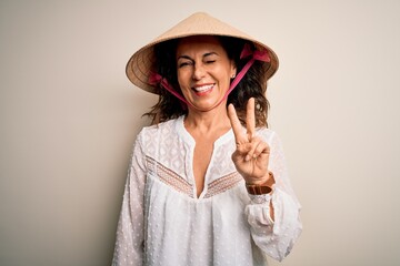 Middle age brunette woman wearing asian traditional conical hat over white background smiling with happy face winking at the camera doing victory sign. Number two.