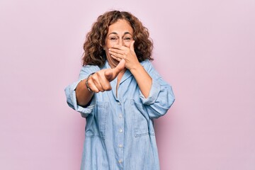Middle age beautiful woman wearing casual denim shirt standing over pink background laughing at you, pointing finger to the camera with hand over mouth, shame expression